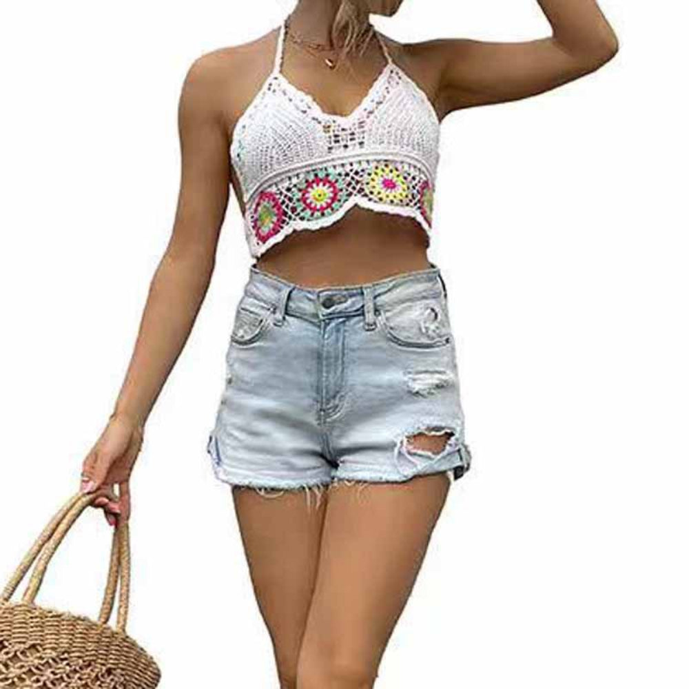 A woman wearing a white crochet halter crop top with delicate openwork lace and colorful floral granny square motifs along the hem. Paired with light-wash distressed denim shorts and a woven straw tote bag, showcasing a boho, summery, and feminine style.
