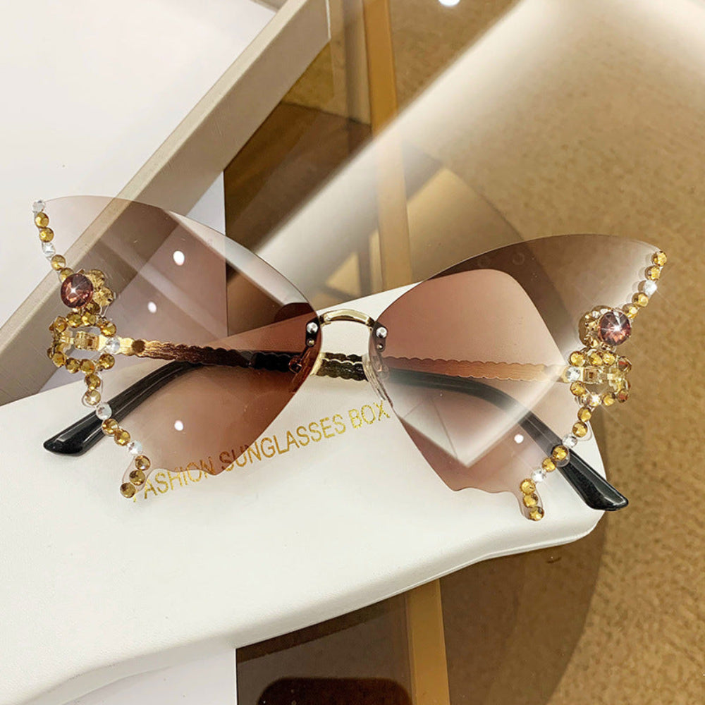 Rimless butterfly sunglasses with brown gradient lenses and gold crystal wing details, displayed on a white glasses case.