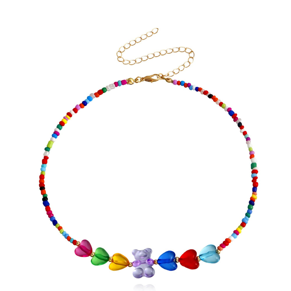 Rainbow beaded choker necklace with multicolored seed beads and large translucent heart-shaped beads in pink, green, yellow, blue, red, and light blue. Features a central purple gummy bear charm and silver lobster clasp with extender chain. Fun, vibrant statement piece displayed on a clean white background.