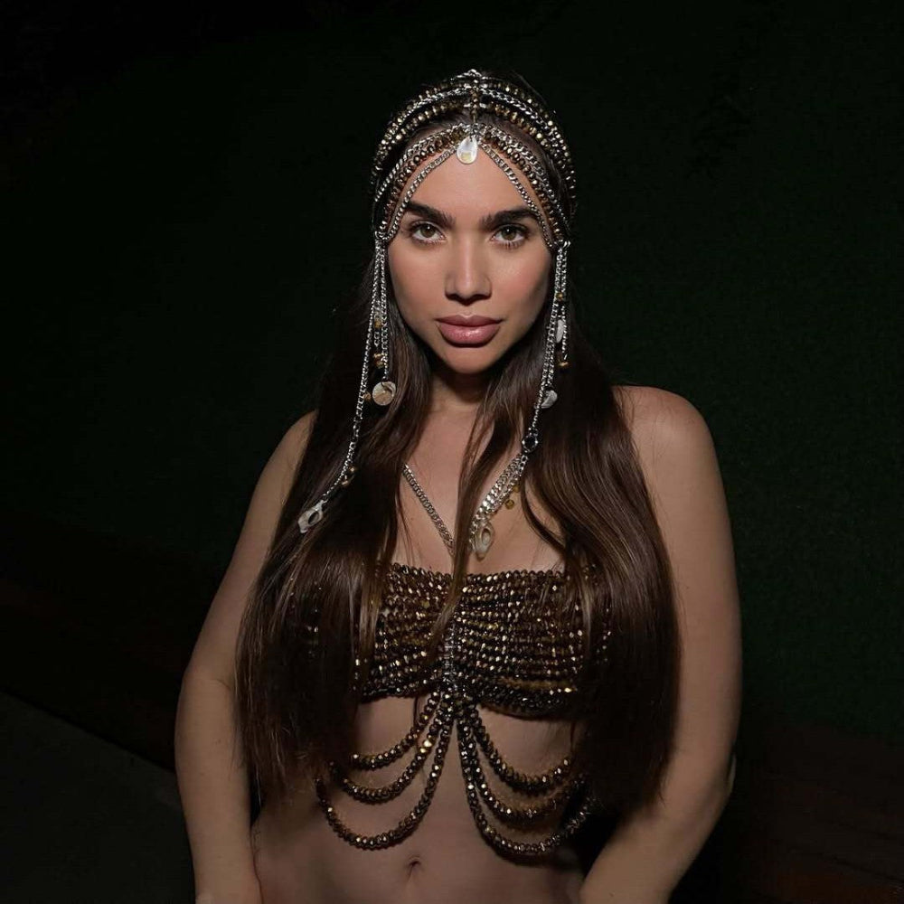 Woman with long brown hair wearing a luxurious gold beaded chainmail crop top with cascading fringe chains, paired with an ornate silver and gold chain headpiece featuring dangling coins and charms. Dramatic studio portrait against a dark background.