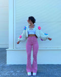 Woman with dark hair posing confidently in a white knit cardigan with colorful pom-poms, white crop top, pink pants, and white sneakers, standing in front of a white slatted wall.