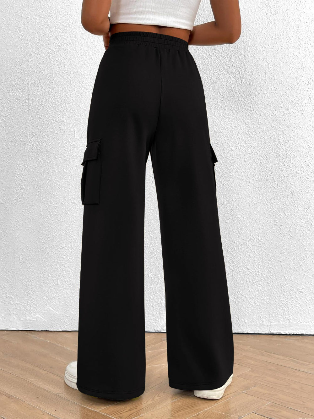 Woman wearing black wide leg cargo pants with side pockets shown from the rear paired with a white top and white sneakers against a textured white wall background