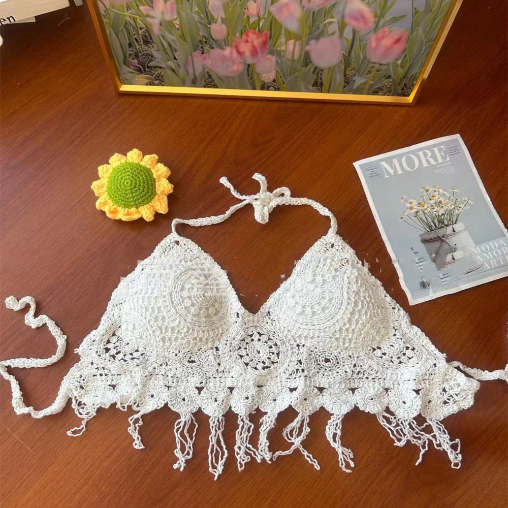 A white handmade crocheted bikini top with intricate lace detailing, triangle cups, halter neck ties, and a delicate fringed hem, displayed flat on a wooden surface. To the left is a small yellow crocheted sunflower, and to the right is a magazine titled "MORE". In the background, a framed painting of pink tulips is visible.