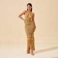 Woman posing in mustard yellow crochet halter maxi dress with fringe hem, deep plunge neckline, and waist cutouts against beige background.