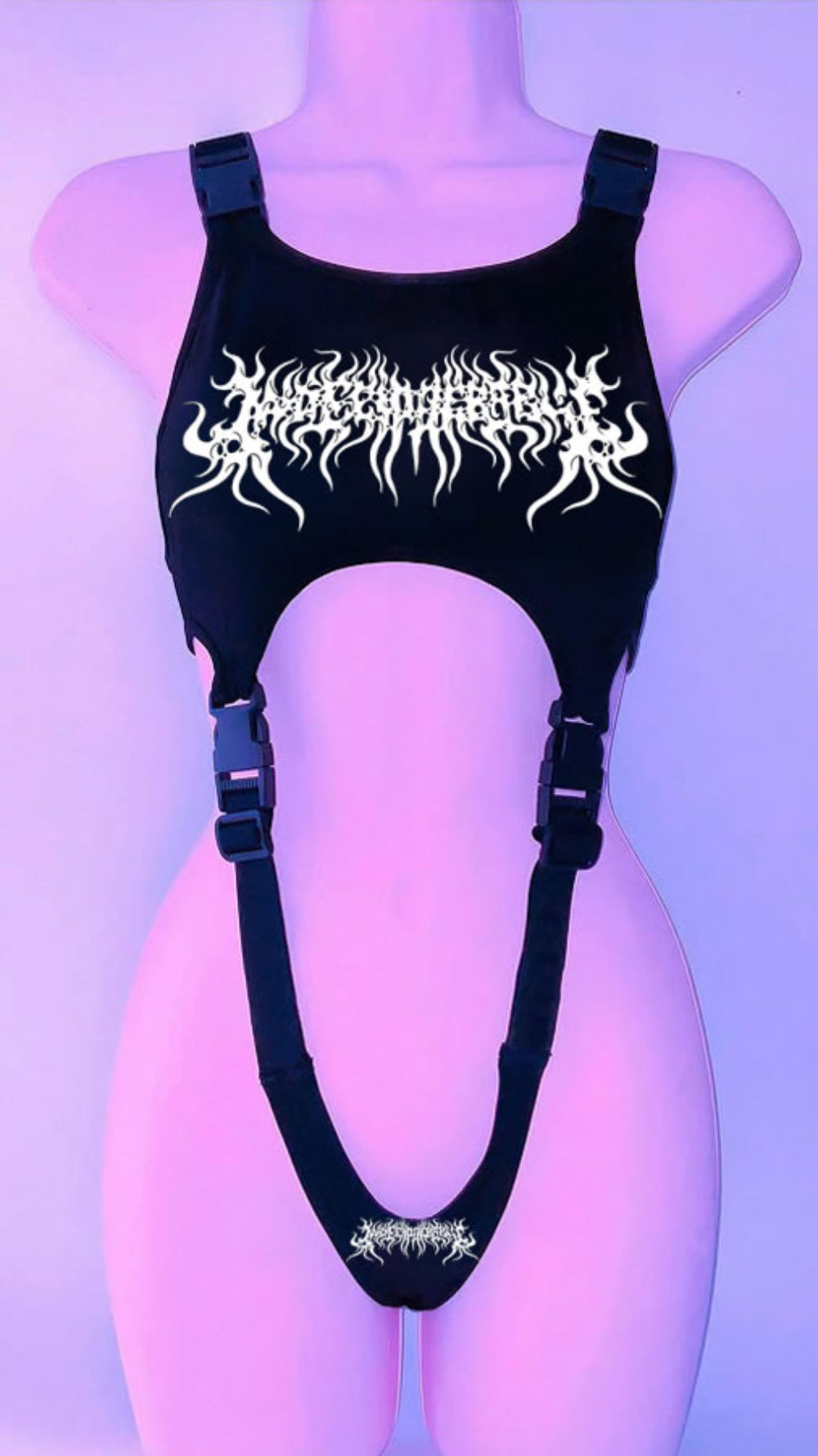 Black harness-style crop top on a mannequin with adjustable buckle straps and side cut-outs. Features a bold white graphic print with skull and stylized text on the chest, plus a smaller matching graphic at the bottom, shown under purple lighting.