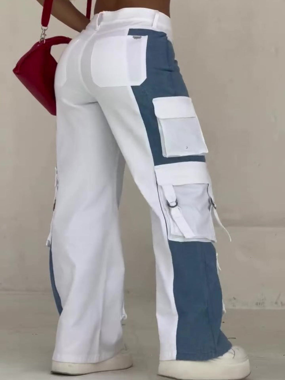 Back and side view of a woman wearing high-waisted color-block cargo jeans with white and blue denim panels, featuring multiple large flap pockets, metal hardware, and D-rings. Styled with a white crop top, chunky white platform sneakers, and a small red crossbody bag