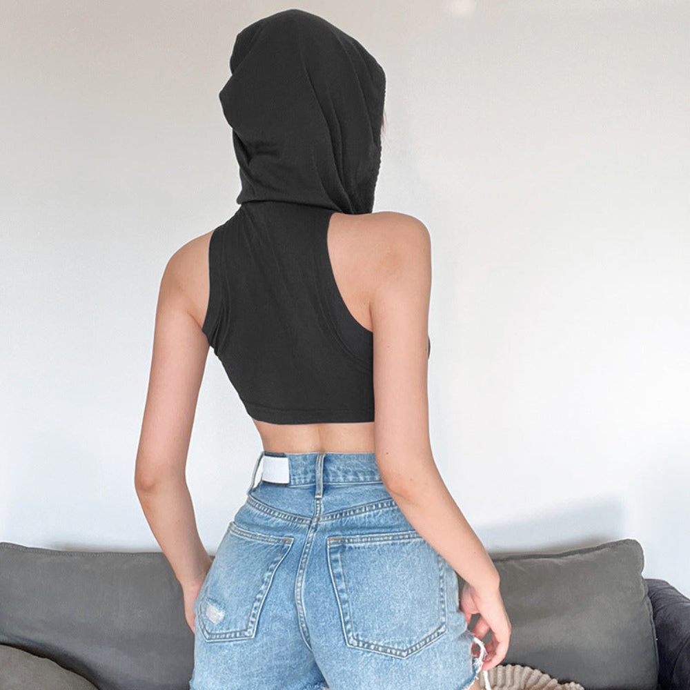 Back view of a woman wearing a black sleeveless hooded crop top with the hood pulled up over her head. Paired with light-wash distressed blue denim jeans, posing with hands near her back pockets in front of a gray couch.