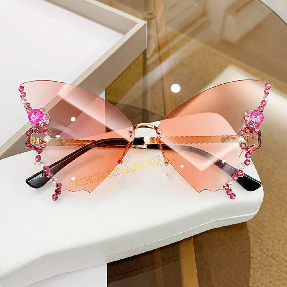 Rimless butterfly sunglasses with peach-pink lenses and pink crystal wing details, displayed on a white glasses case.
