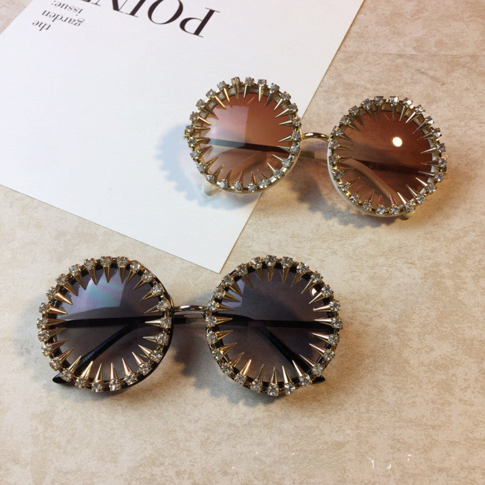 Round steampunk sunglasses with spiked rhinestone rims in gold and black, styled on a beige surface.