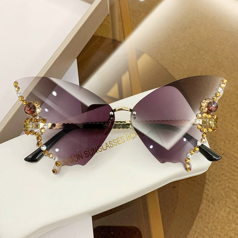 Rimless butterfly sunglasses with brown-purple gradient lenses and gold crystal wing details, displayed on a white glasses case.