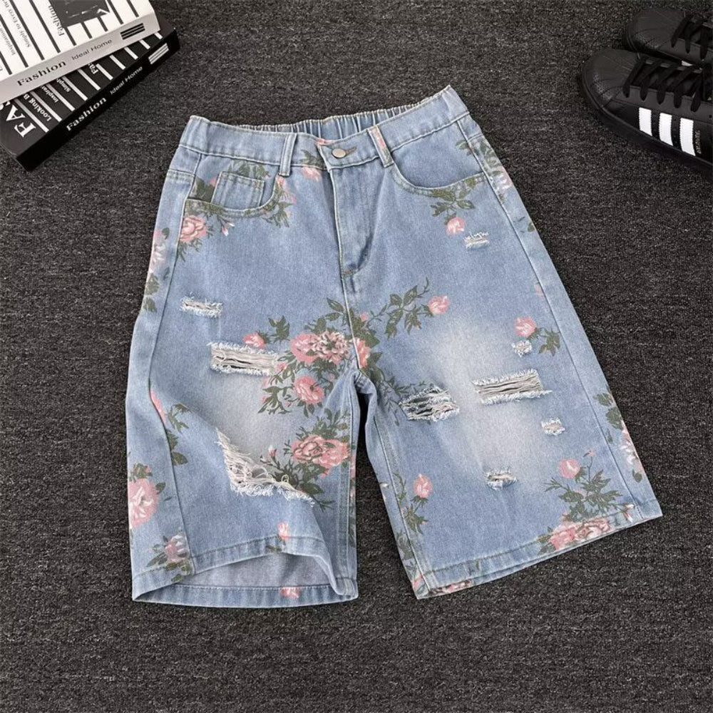 Light blue distressed denim shorts with pink floral rose prints, frayed rips, and raw hems, styled flat on a dark surface with black sneakers and a fashion book. Vintage-inspired product photography for Boutique Jolie.