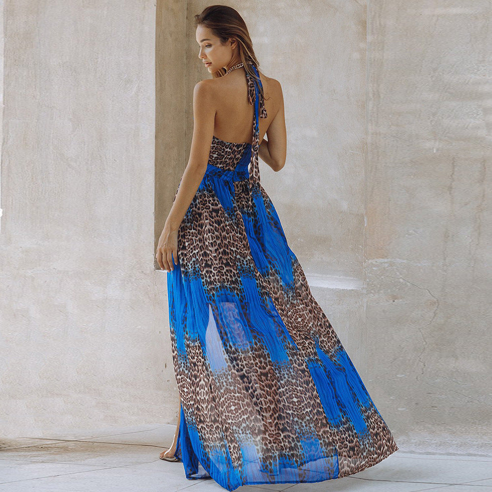 Model shown from behind wearing a stunning long halter maxi dress featuring bold leopard print mixed with vibrant blue abstract panels and pleated sheer fabric. Elegant back view highlighting the tie closure and flowing silhouette, posed gracefully in a minimalist stone architectural setting.