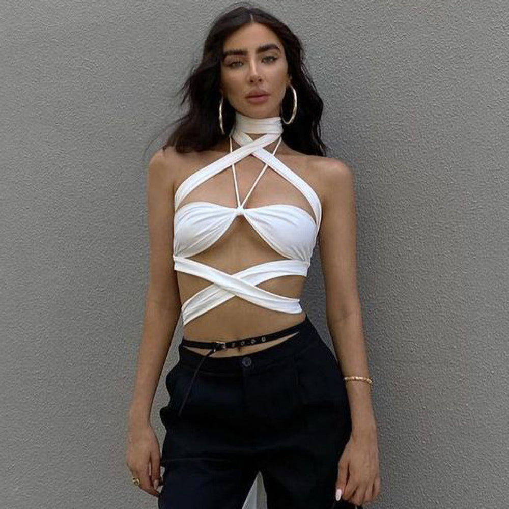 Alt text: A confident woman with long, dark wavy hair wearing a white halter crop top with dramatic crisscross straps, front cutouts, and wrapped bands around the midriff. She is styled with black trousers, a thin belt, large hoop earrings, and gold jewelry, posing against a textured gray wall.