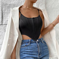 Woman wearing a black corset-style bodysuit with front zipper closure and structured boning, layered under an open white blazer with pearl buttons. Paired with blue denim jeans and gold chain necklace plus bracelet, posing against a textured white wall.
