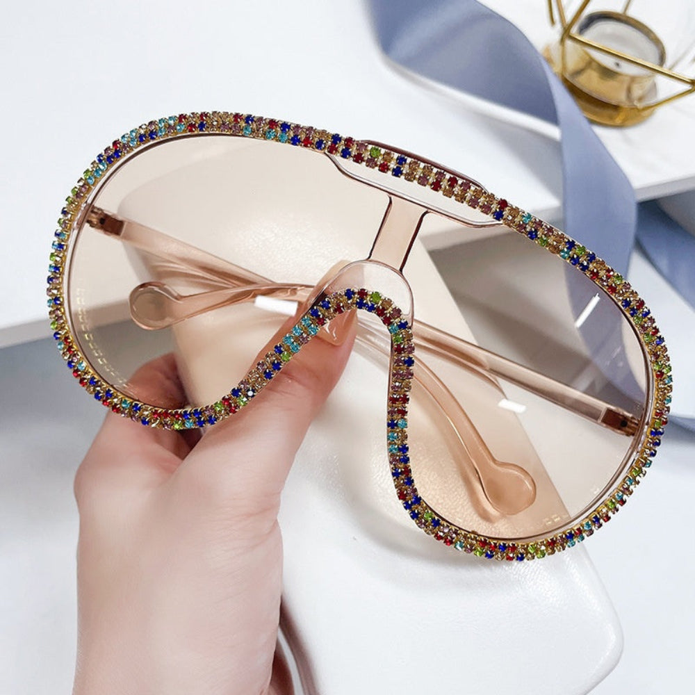 Hand holding oversized champagne-lens shield sunglasses with rainbow rhinestone trim.