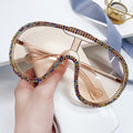 Hand holding oversized champagne-lens shield sunglasses with rainbow rhinestone trim.