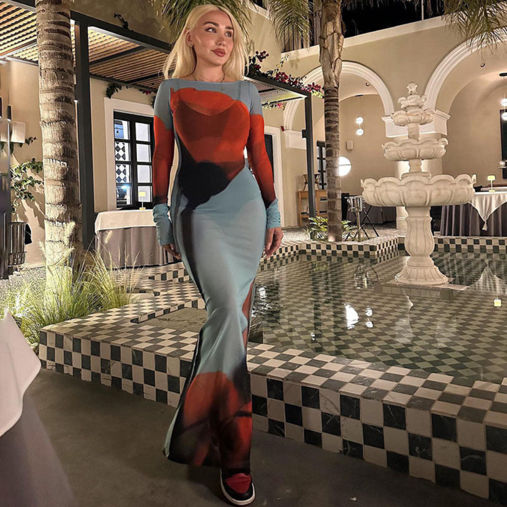 Blonde woman modeling a colorful abstract long dress in red, black, and light blue tones, posing elegantly by a poolside fountain in a stylish outdoor courtyard setting.