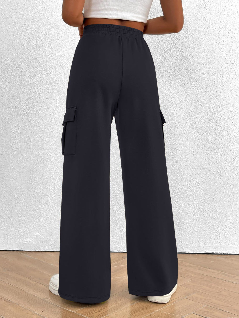 Woman wearing black wide leg cargo pants with side pockets shown from the rear paired with a white top and white sneakers against a textured white wall background