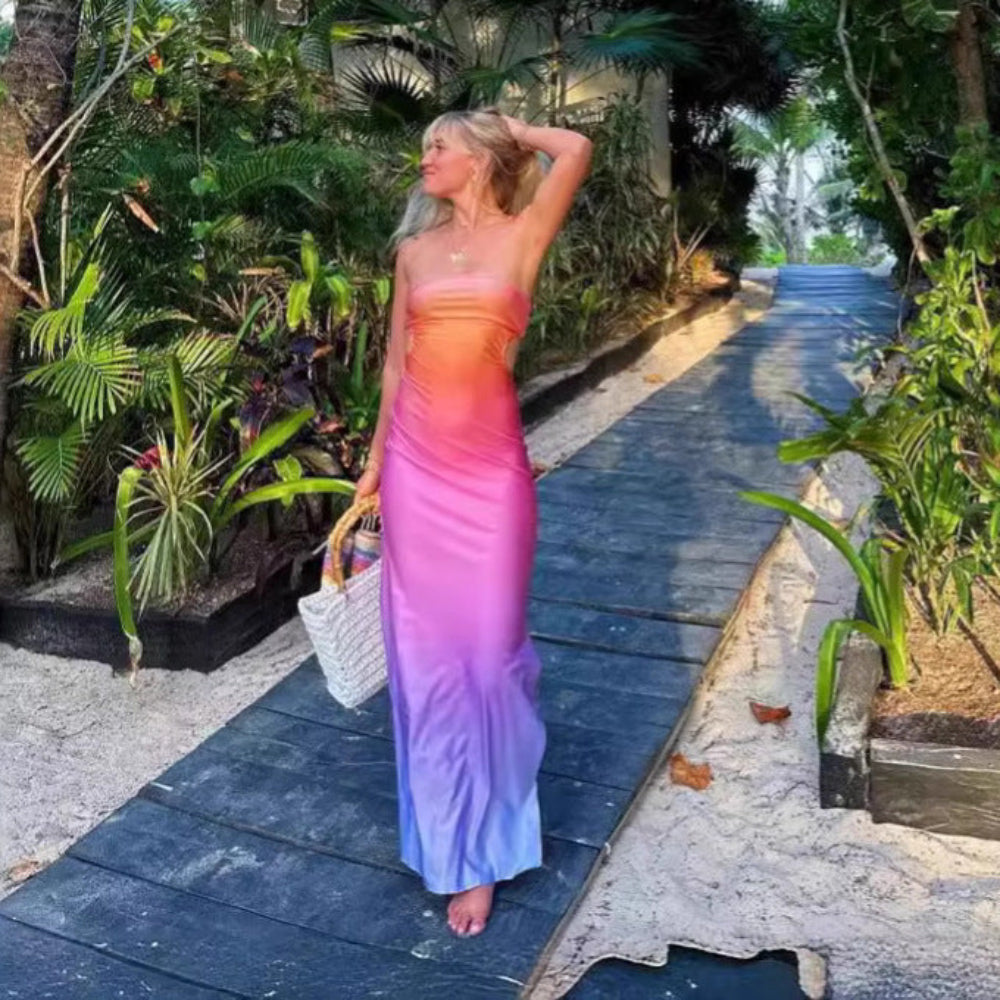 Model in a stunning ombre strapless floor-length dress transitioning from vibrant orange-pink at the top to soft purple at the hem. Side/rear view as she walks along a tropical garden path with dense greenery, one hand touching her hair and holding a woven tote bag.
