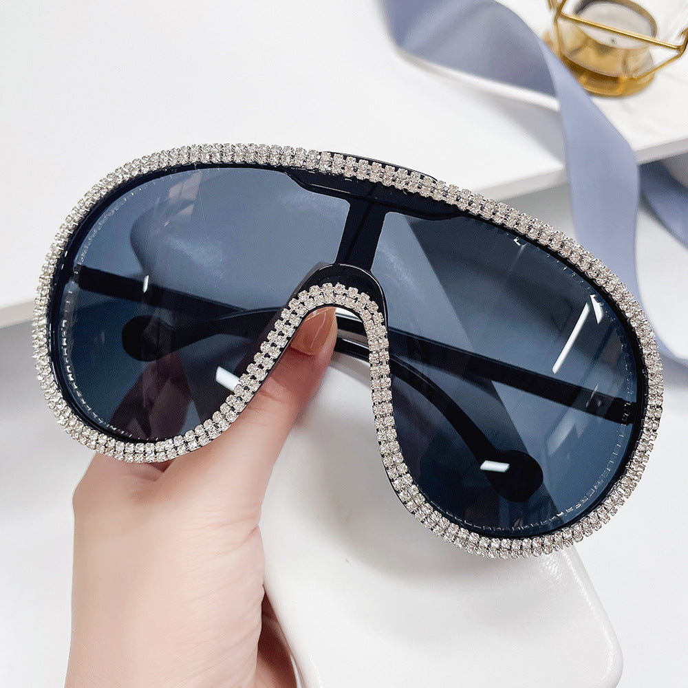 Hand holding oversized dark-lens shield sunglasses with crystal rhinestone trim.