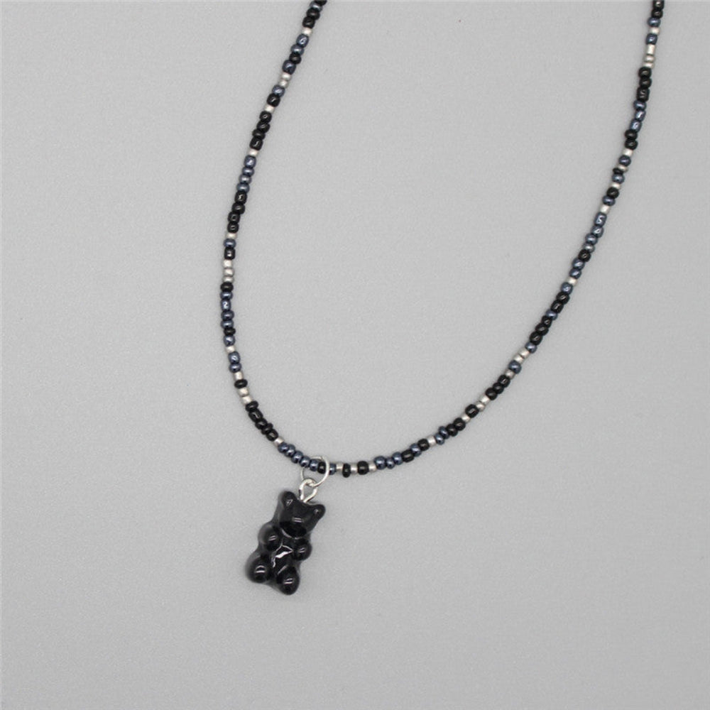 Dark-themed beaded necklace with a mix of black, navy blue, and silver/white seed beads paired with a translucent black gummy bear pendant. Sleek single-strand design laid flat on a light gray background, offering a bold, edgy, and playful look.